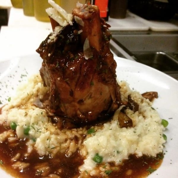 Braised lamb shank with rich brown sauce, creamy risotto, peas, and shaved cheese on a white plate.