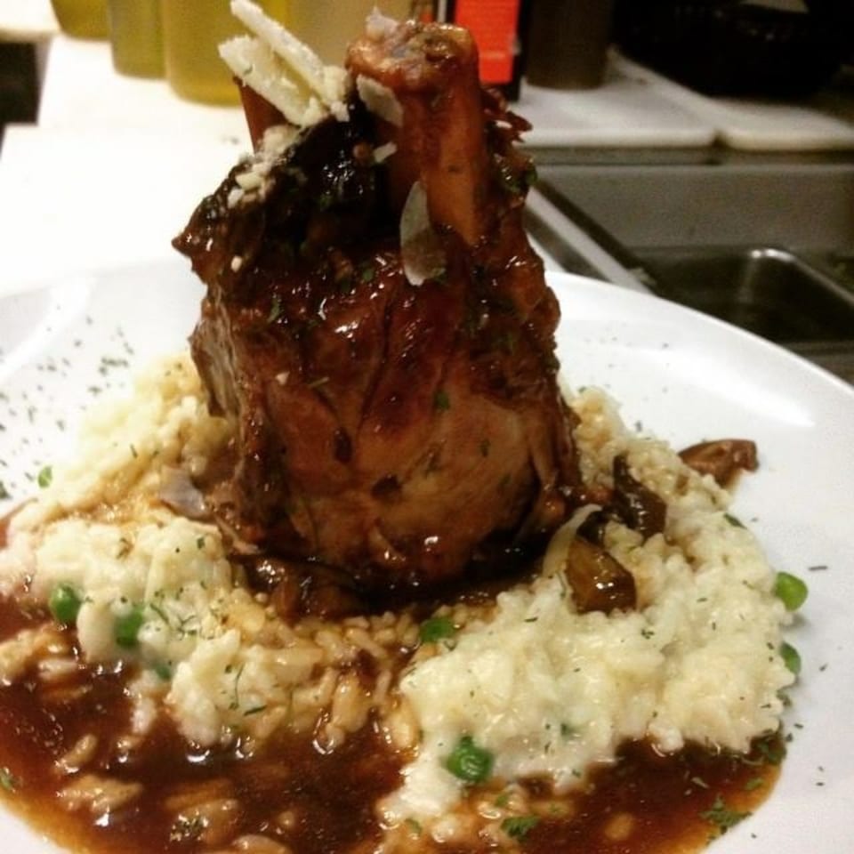 Braised lamb shank with rich brown sauce, creamy risotto, peas, and shaved cheese on a white plate.