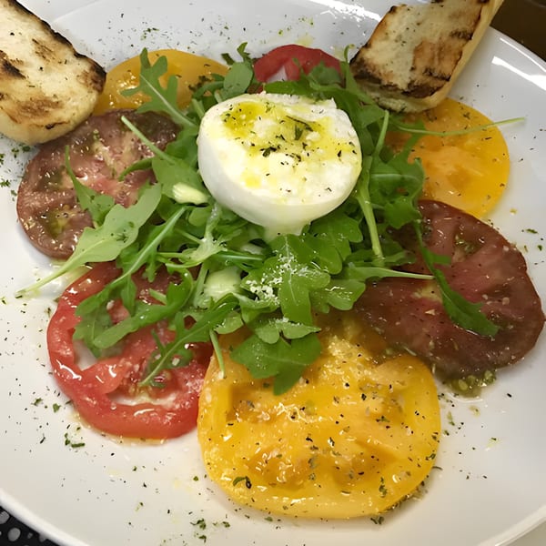 Heirloom tomato salad with burrata, arugula, olive oil, and grilled bread on a white plate.