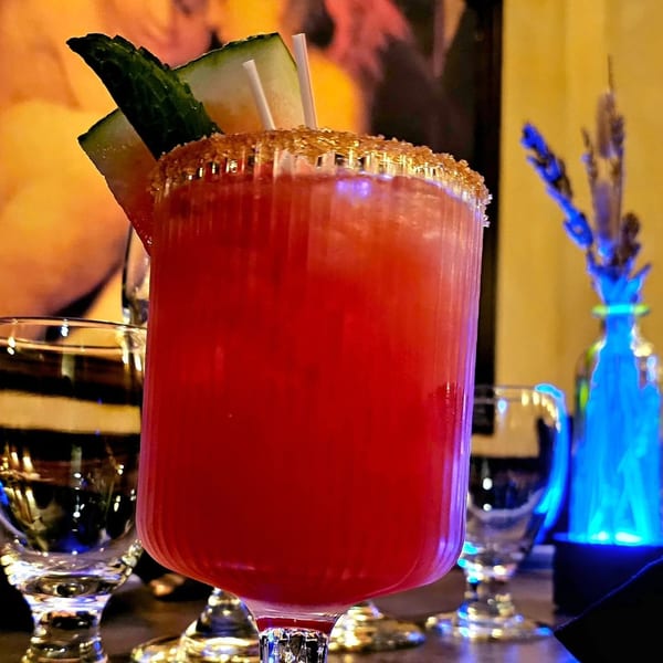 Red cocktail in a stemmed glass, garnished with cucumber and lime, on a warmly lit restaurant bar.