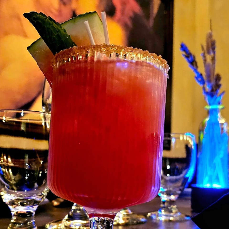 Red cocktail in a stemmed glass, garnished with cucumber and lime, on a warmly lit restaurant bar.